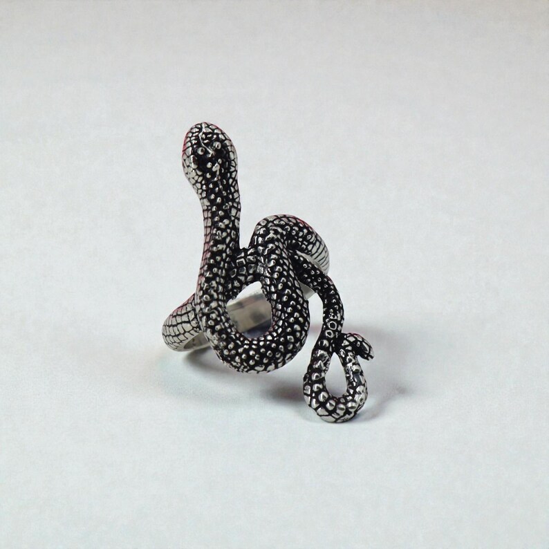 May include: A silver snake ring with a detailed, textured pattern. The snake is coiled, with its head raised, and the body wraps around the finger. The ring has a polished finish and is set against a white backdrop.
