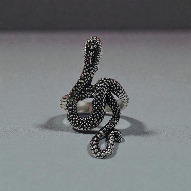 May include: A silver-coloured ring in the shape of a snake, coiled and upright. The snake's body is detailed with textured scales, and its head is raised. The ring is set against a grey background.