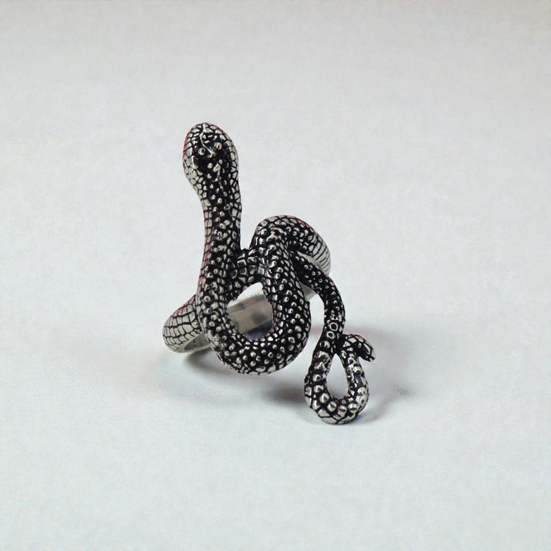 May include: A silver snake ring with a detailed, textured pattern. The snake is coiled, with its head raised, and the body wraps around the finger. The ring has a polished finish and is set against a white backdrop.