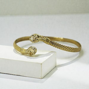 May include: A gold-coloured snake bracelet. The bracelet has a snake head design on each end, with detailed scales and patterns. The bracelet is open, allowing it to be adjusted to fit different wrist sizes. The snake heads are facing opposite directions.