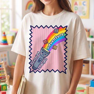 Op de afbeelding: Een crèmekleurig t-shirt met een afbeelding van een lachende zaklamp die een regenboogstraal uitstraalt met zwevende alfabetblokken. De tekst luidt Learning is a Bright adventure. Het ontwerp is omlijst door een donkerblauwe gekartelde rand.