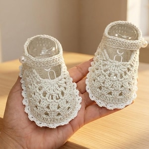 May include: A pair of cream-colored, crocheted baby shoes. The shoes feature a delicate openwork design, a Mary Jane strap with a pearl accent, and a scalloped edge. The shoes are designed to fit a baby's foot.