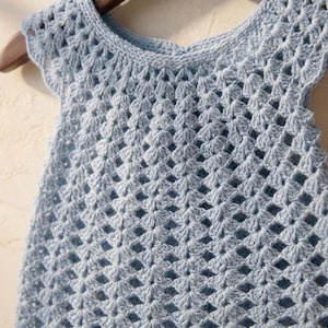 May include: A light blue, handmade crochet top with a delicate, openwork pattern. The sleeveless top features a scalloped neckline and armholes. The garment is displayed on a wooden hanger, showcasing the intricate stitchwork and airy design.