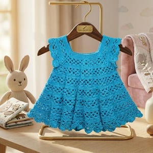 May include: A bright turquoise crochet baby dress with a scalloped hem and cap sleeves hangs on a wooden hanger. A stuffed bunny and a baby book are on a wooden table. The dress is a handmade item.