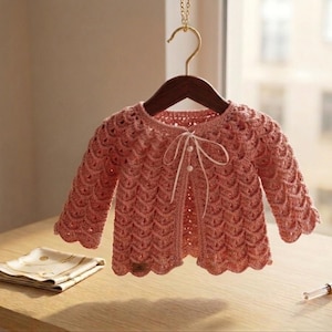 May include: A coral-coloured, crocheted cardigan with a scalloped hem and sleeves hangs on a wooden hanger. The open-front sweater features a delicate, textured stitch pattern and a white ribbon tie closure. A folded napkin and perfume bottle are on the table.