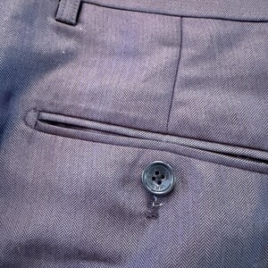 May include: Close-up of dark gray dress pants with a herringbone pattern. The pants feature a buttoned back pocket and a button with the word "VERSACE" on it. The fabric has a subtle sheen.