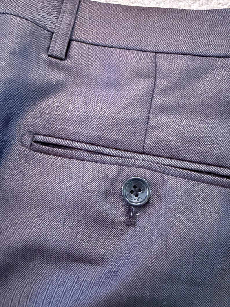 May include: Close-up of dark gray dress pants with a herringbone pattern. The pants feature a buttoned back pocket and a button with the word "VERSACE" on it. The fabric has a subtle sheen.
