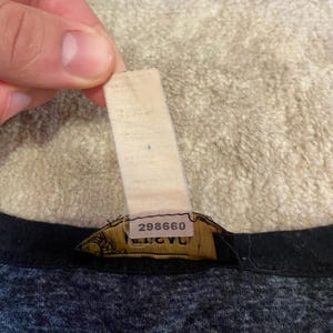 May include: Close-up of a garment tag with the number "298660" printed on it. The tag is attached to a dark gray and black fabric. The tag is beige and has some text printed on it.