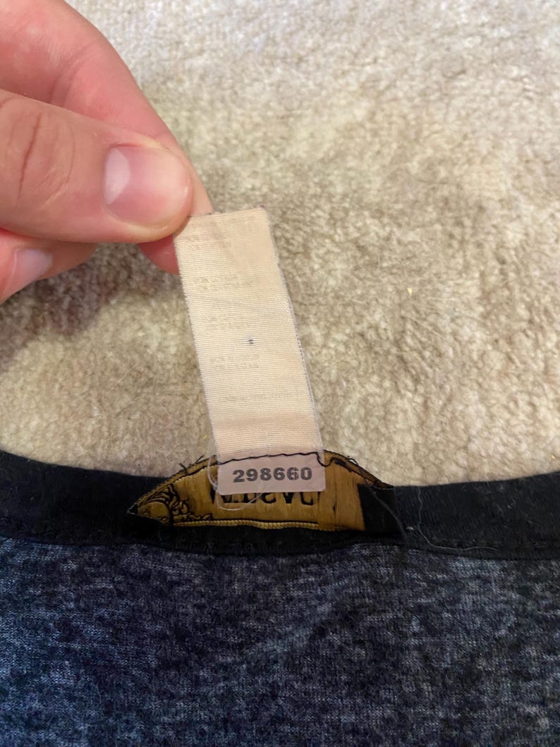 May include: Close-up of a garment tag with the number "298660" printed on it. The tag is attached to a dark gray and black fabric. The tag is beige and has some text printed on it.