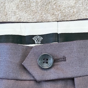 May include: Close-up of a pair of gray herringbone-patterned trousers. The button is dark gray with the word VERSACE around the edge. The label features the Versace logo.