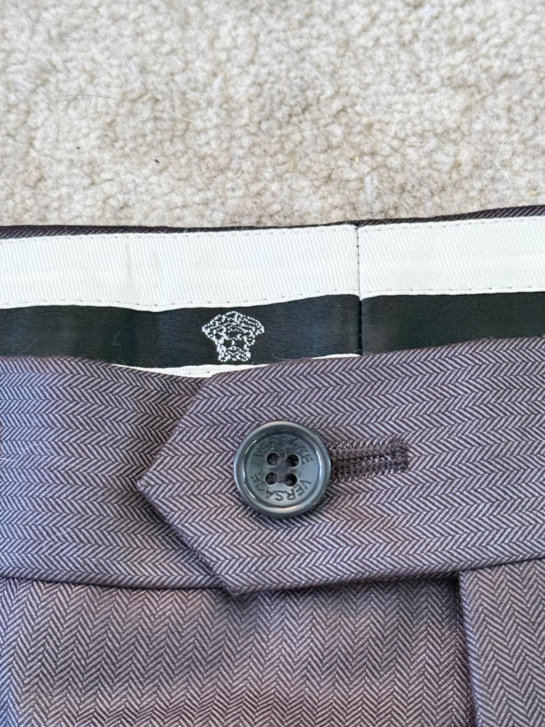 May include: Close-up of a pair of gray herringbone-patterned trousers. The button is dark gray with the word VERSACE around the edge. The label features the Versace logo.