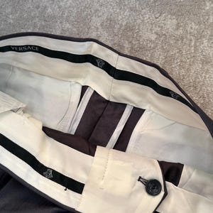 May include: Close-up of dark gray dress pants with a white waistband and black accents. The waistband features the brand name "VERSACE" and a decorative emblem. The pants have a button closure and a herringbone pattern.