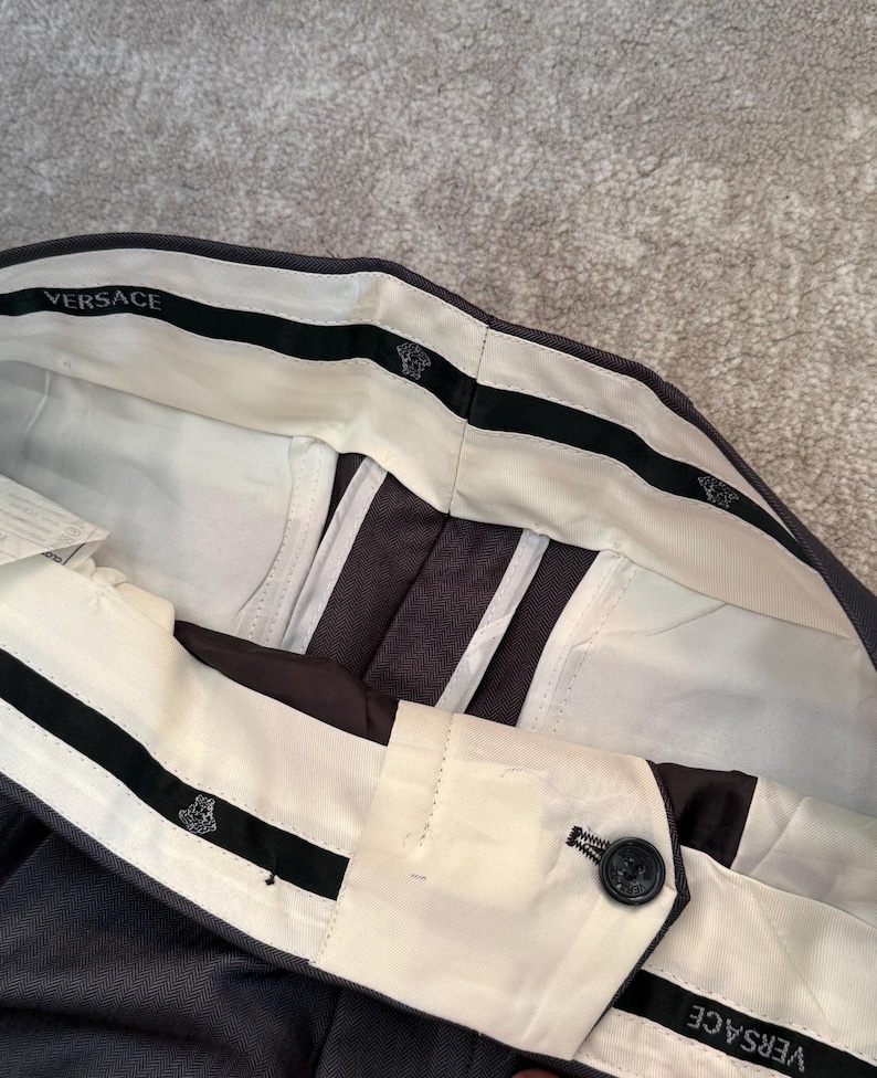 May include: Close-up of dark gray dress pants with a white waistband and black accents. The waistband features the brand name "VERSACE" and a decorative emblem. The pants have a button closure and a herringbone pattern.