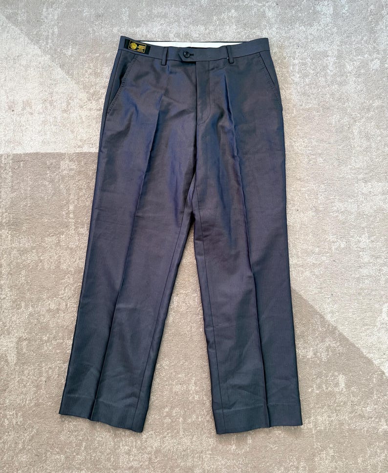 May include: Dark gray dress pants with a button closure and pleats. The pants have two front pockets and two back pockets. A small tag is visible on the waistband. The pants are laid flat on a light-colored surface.