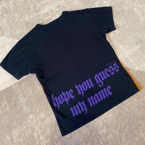 May include: Black t-shirt with short sleeves. The back of the shirt features the phrase "hope you guess my name" in purple gothic lettering. An orange stripe is visible on the right side of the shirt.