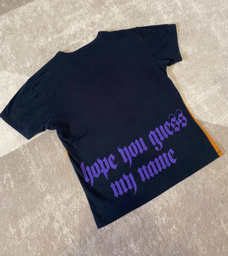 May include: Black t-shirt with short sleeves. The back of the shirt features the phrase "hope you guess my name" in purple gothic lettering. An orange stripe is visible on the right side of the shirt.