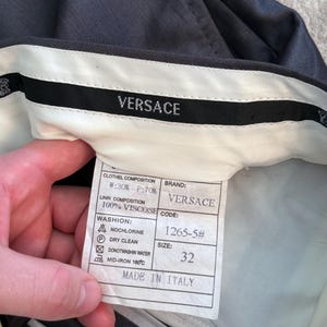 May include: Dark gray Versace trousers with a white interior waistband. The label shows the brand name, size 32, and fabric composition. The label also includes washing instructions and the text "MADE IN ITALY".