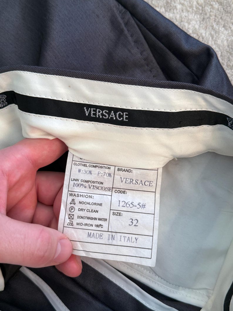May include: Dark gray Versace trousers with a white interior waistband. The label shows the brand name, size 32, and fabric composition. The label also includes washing instructions and the text "MADE IN ITALY".