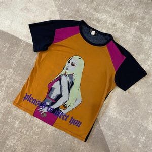 May include: Mustard yellow t-shirt with navy blue and magenta color block sleeves. The shirt features a graphic of a woman with long blonde hair and the text "please to meet you" in purple.