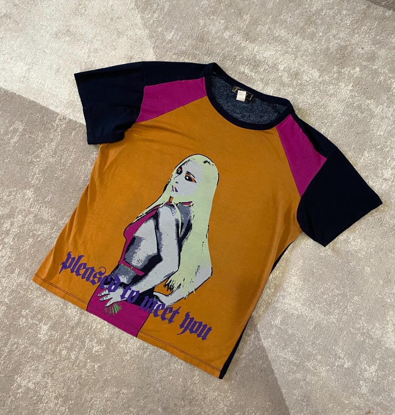 May include: Mustard yellow t-shirt with navy blue and magenta color block sleeves. The shirt features a graphic of a woman with long blonde hair and the text "please to meet you" in purple.