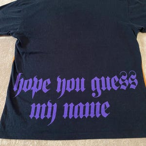 May include: Black t-shirt with the phrase "hope you guess my name" in purple gothic font. The shirt is made of a soft material and has short sleeves. The shirt is laying flat on a neutral surface.
