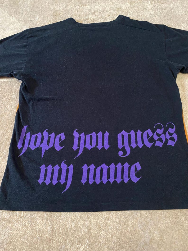 May include: Black t-shirt with the phrase "hope you guess my name" in purple gothic font. The shirt is made of a soft material and has short sleeves. The shirt is laying flat on a neutral surface.