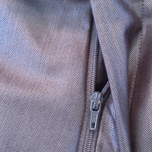 May include: Close-up of a section of dark gray fabric with a herringbone pattern. A silver zipper is partially open, revealing the fabric's texture and weave. The fabric appears to be a tailored material, possibly for a garment.