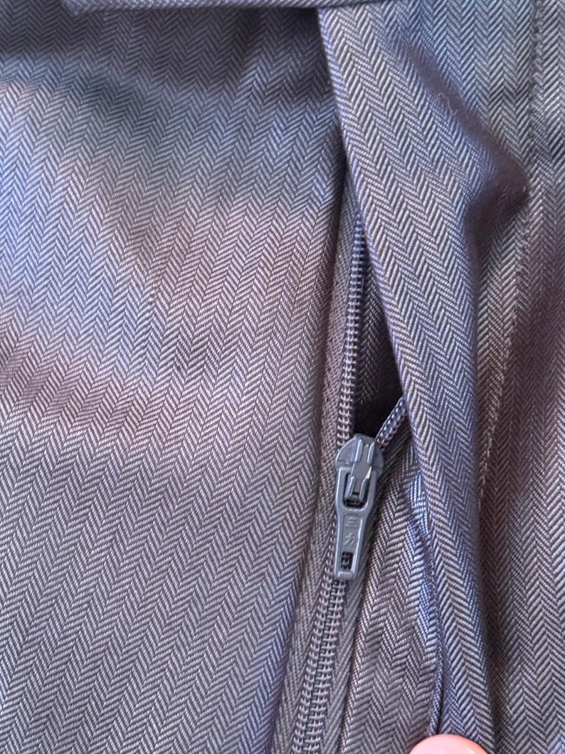 May include: Close-up of a section of dark gray fabric with a herringbone pattern. A silver zipper is partially open, revealing the fabric's texture and weave. The fabric appears to be a tailored material, possibly for a garment.