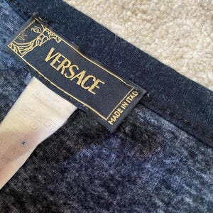 May include: Close-up of a navy blue garment with a black trim. A rectangular label with the gold-colored text "VERSACE" and "MADE IN ITALY" is attached to the garment. The label has a gold emblem.
