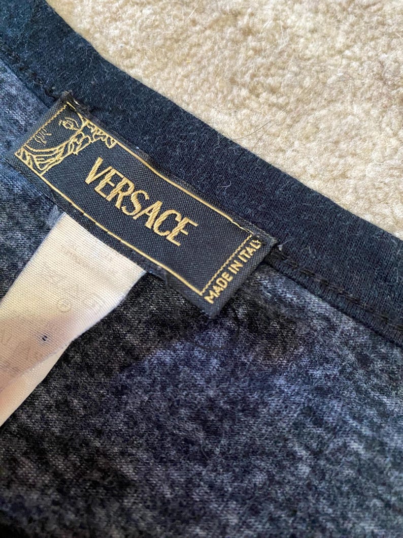 May include: Close-up of a navy blue garment with a black trim. A rectangular label with the gold-colored text "VERSACE" and "MADE IN ITALY" is attached to the garment. The label has a gold emblem.