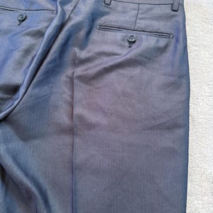 May include: Close-up of dark gray dress pants. The pants have two back pockets with button closures and a button and zipper closure at the front. The fabric has a subtle texture.