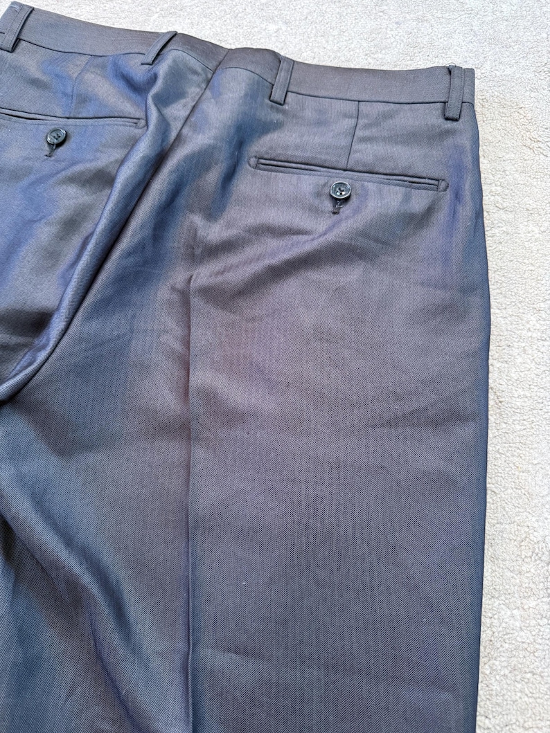 May include: Close-up of dark gray dress pants. The pants have two back pockets with button closures and a button and zipper closure at the front. The fabric has a subtle texture.