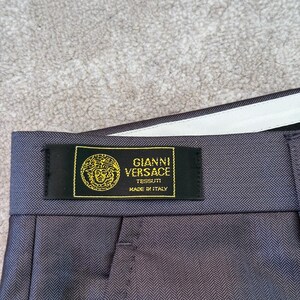 May include: Close-up of gray herringbone-patterned trousers with a black label. The label reads "GIANNI VERSACE TESSUTI MADE IN ITALY" in gold lettering. The trousers have a white waistband.