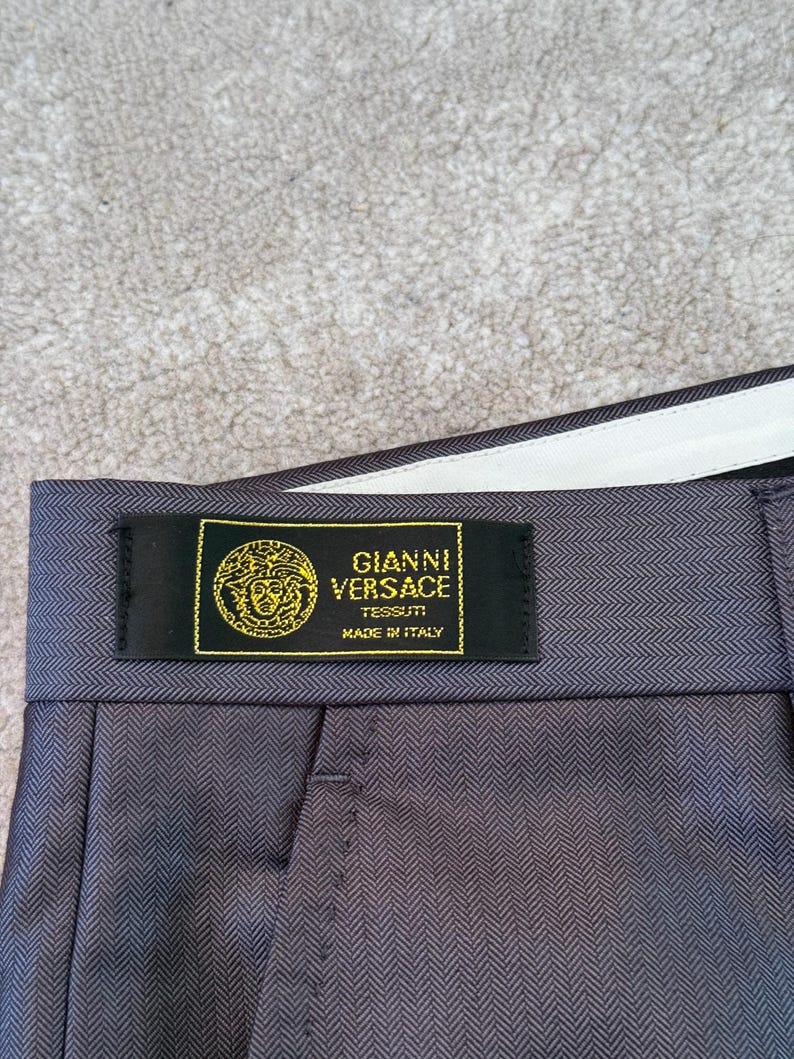 May include: Close-up of gray herringbone-patterned trousers with a black label. The label reads "GIANNI VERSACE TESSUTI MADE IN ITALY" in gold lettering. The trousers have a white waistband.