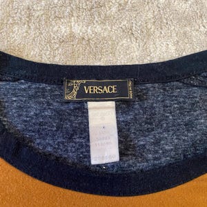 May include: Close-up of a t-shirt with a black neckline and a mustard yellow and navy blue color block design. The shirt features a Versace label with the brand name and logo.