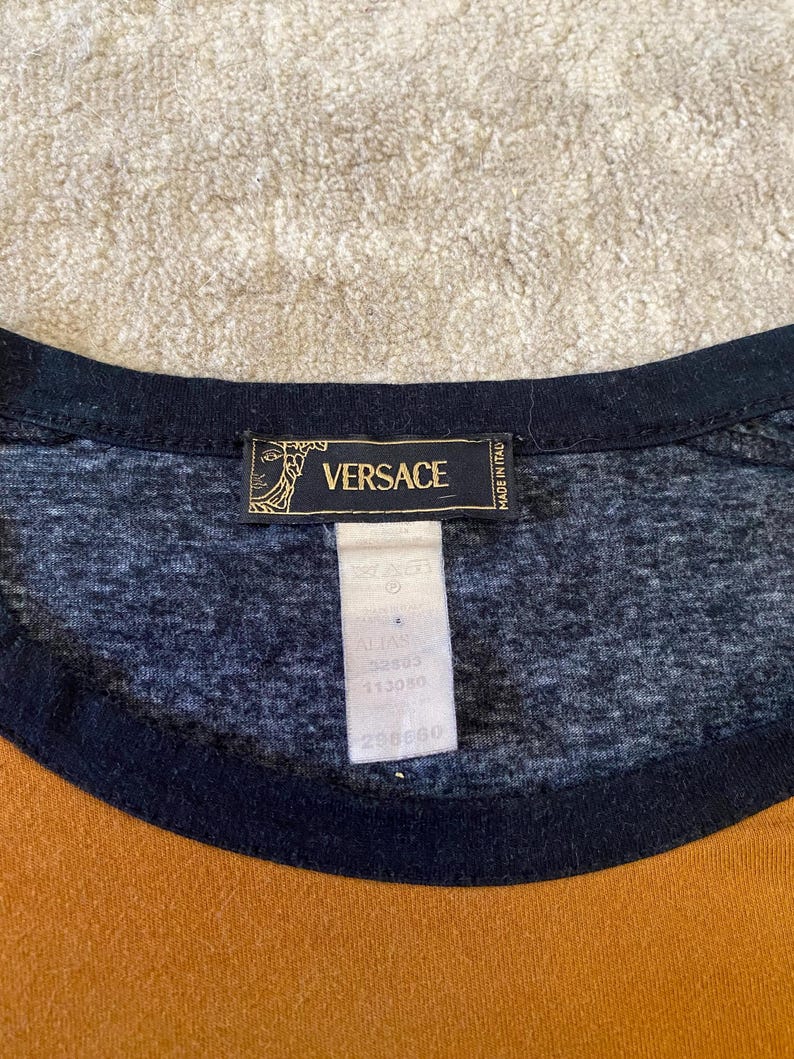 May include: Close-up of a t-shirt with a black neckline and a mustard yellow and navy blue color block design. The shirt features a Versace label with the brand name and logo.