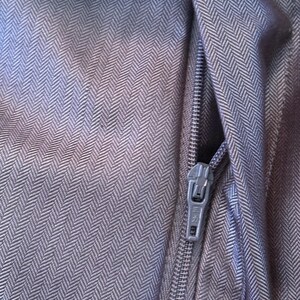 May include: Close-up of a section of dark gray fabric with a herringbone pattern. A silver zipper is partially open, revealing the interior. The fabric has a subtle sheen, and the zipper pull is visible.
