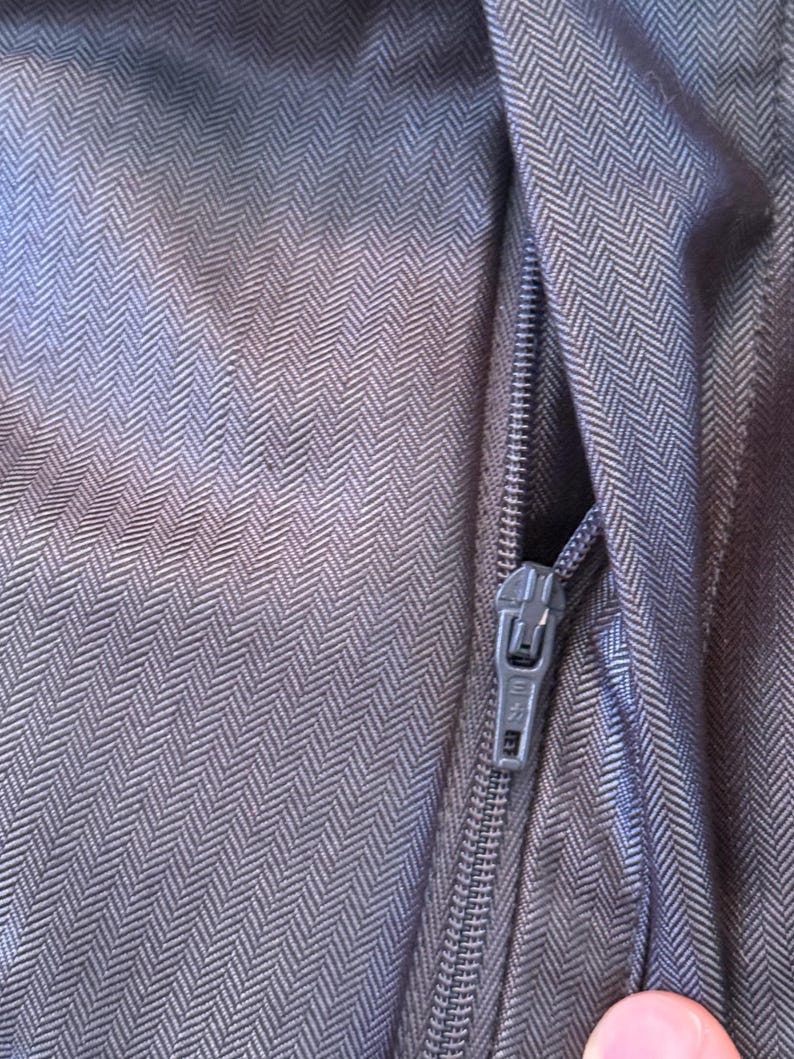 May include: Close-up of a section of dark gray fabric with a herringbone pattern. A silver zipper is partially open, revealing the interior. The fabric has a subtle sheen, and the zipper pull is visible.