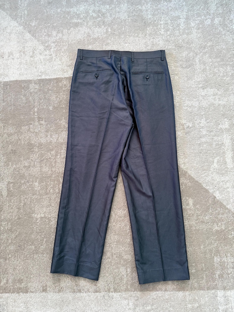 May include: Dark gray dress pants with two back pockets and button closures. The pants are made of a smooth fabric and have a classic, tailored design. The pants are laid flat on a light gray surface.