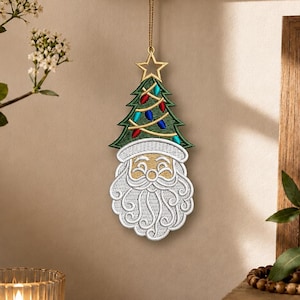May include: A hanging Christmas ornament featuring an embroidered Santa Claus face with a white beard and mustache, topped with a green Christmas tree decorated with colorful lights and a gold star, suspended by a thin gold cord from a hook.