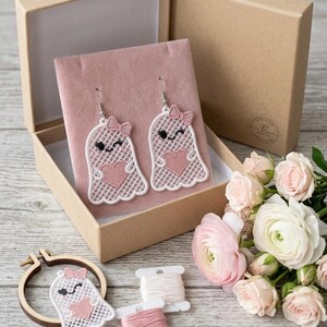 May include: A pair of white lace ghost earrings featuring pink heart accents and small pink bows, displayed on a pink velvet insert inside a cardboard box. An additional matching ghost patch, two spools of embroidery thread, and pink roses are included.