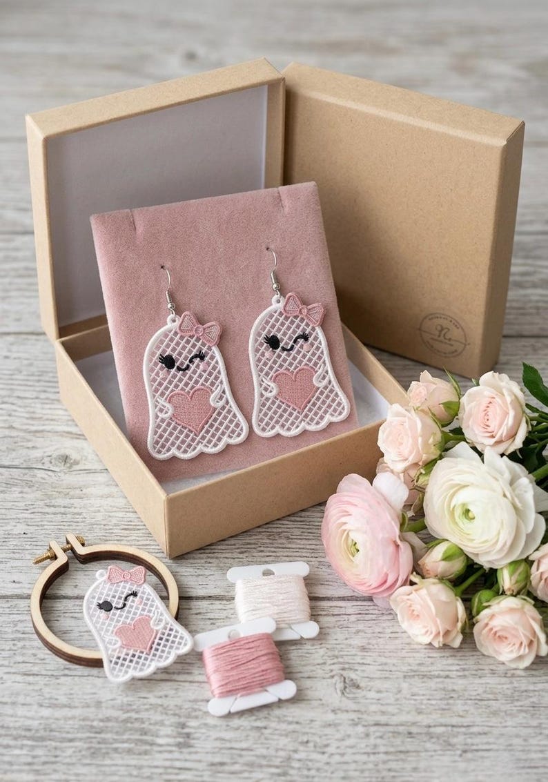 May include: A pair of white lace ghost earrings featuring pink heart accents and small pink bows, displayed on a pink velvet insert inside a cardboard box. An additional matching ghost patch, two spools of embroidery thread, and pink roses are included.