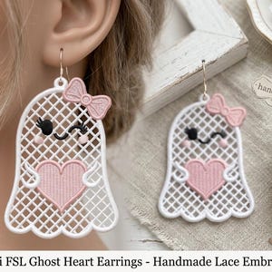 May include: A pair of white ghost-shaped earrings featuring a lattice pattern, a pink embroidered heart, and a small pink bow. The ghost has black embroidered eyes with eyelashes and pink cheeks. Each earring hangs from a silver-toned metal hook.