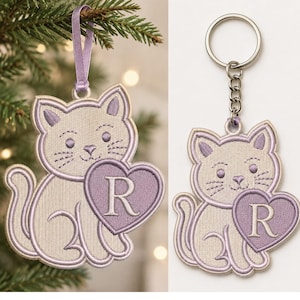 May include: An embroidered cat ornament and keychain set featuring a light beige cat with lavender accents. The cat holds a lavender heart with the letter R stitched in the center. The ornament includes a lavender ribbon loop for hanging.