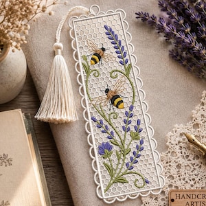May include: A rectangular white lace bookmark featuring an intricate honeycomb pattern, embroidered with two yellow and black bumblebees and purple lavender sprigs. The bookmark is finished with a scalloped edge and a cream-colored fabric tassel.