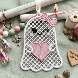May include: A white embroidered ghost ornament with a pink heart and bow, featuring a grid pattern. It is surrounded by pink and green wooden beads, two small glass jars with floral contents, a metal key, two thread spools, and a wooden heart.