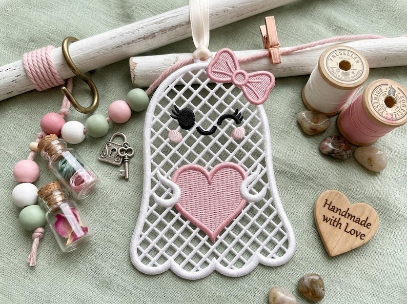 May include: A white embroidered ghost ornament with a pink heart and bow, featuring a grid pattern. It is surrounded by pink and green wooden beads, two small glass jars with floral contents, a metal key, two thread spools, and a wooden heart.