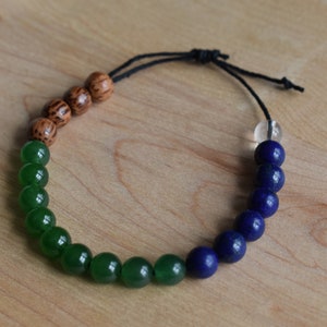 May include: A beaded bracelet featuring a gradient of colors. The bracelet transitions from brown wooden beads to green, then to blue beads, and ends with a clear bead. The bracelet is strung on a black cord.