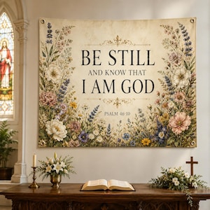Puede incluir: Un tapiz beige con el texto "BE STILL AND KNOW THAT I AM GOD" y "PSALM 46:10", rodeado de ilustraciones florales en rosa, blanco, amarillo y morado. El tapiz está colgado en una iglesia.