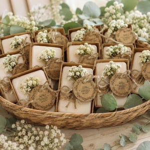Op de afbeelding: Een verzameling rechthoekige beige zeepblokken verpakt in bruine kartonnen doosjes, elk versierd met een takje witte gipskruidbloemen en een rond bruin papieren label vastgebonden met touw, gerangschikt in een ovale mand van gevlochten riet.